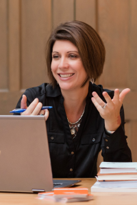 A woman with brown hair sits at a laptop with her hands in the air. She is smiling.