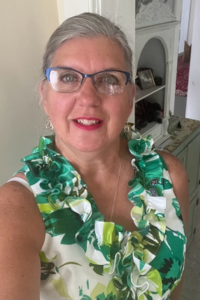 A woman with gray hair that is pulled back takes a selfie. She is wearing glasses and a ruffled, sleeveless, green and white top with a floral pattern. Behind her is a bedroom.