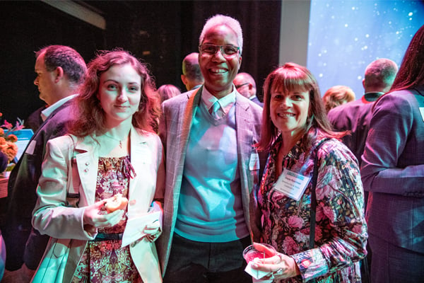 Three people smile during a reception. They are holding drinks. One is a young woman with curly brown hair. Next to her is an older man with white hair. On his other side is a middle-aged woman with brown hair.