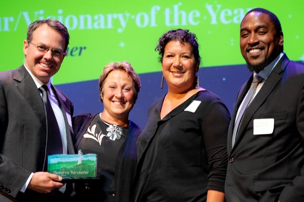 Four people stand in a row smiling. The man on the far left holds an award. Next to him are two women followed by a man. Behind them is a lit screen.