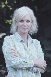 A woman with short white hair stands with her harms folded across her chest. She is lightly smiling at the camera. She is wearing a light-colored button down shirt and behind is a very dark leafy backdground.