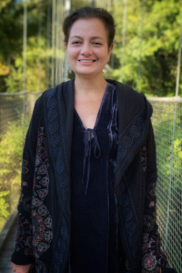 A woman with brown hair that is pulled back stands on a bridge, smiling at the camera. She is wearing a black outfit.