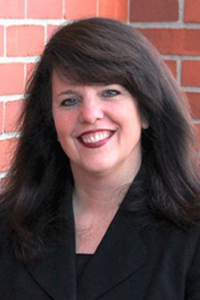 A woman with dark brown hair and red lipstick smiles at the camera. Behind is a red brick wall.