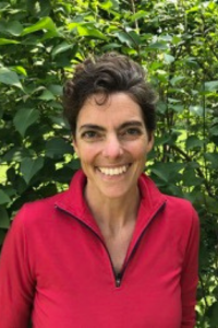 A woman with a pixie cut smiles at the camera. She is wearing a red v-neck top and stands in front of a green leafy hedge.